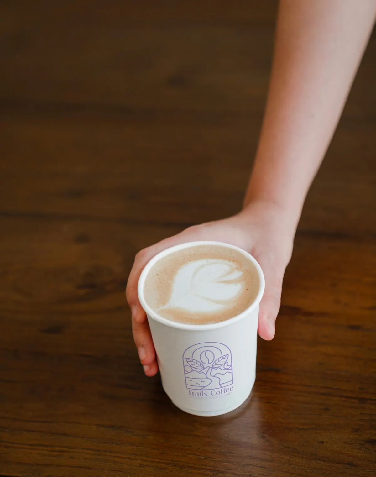 A Trails Coffee latte being handed across a warm wooden table