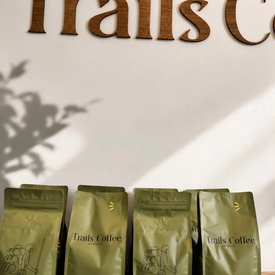 Four green Trails Coffee bags arranged in sunlight beneath the café wordmark